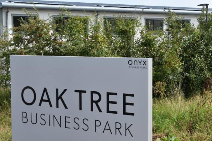 Welcome board at Oak Tree Business Park, Kingskerswell, framed by a row of bushes in the foreground, with industrial units in the background.