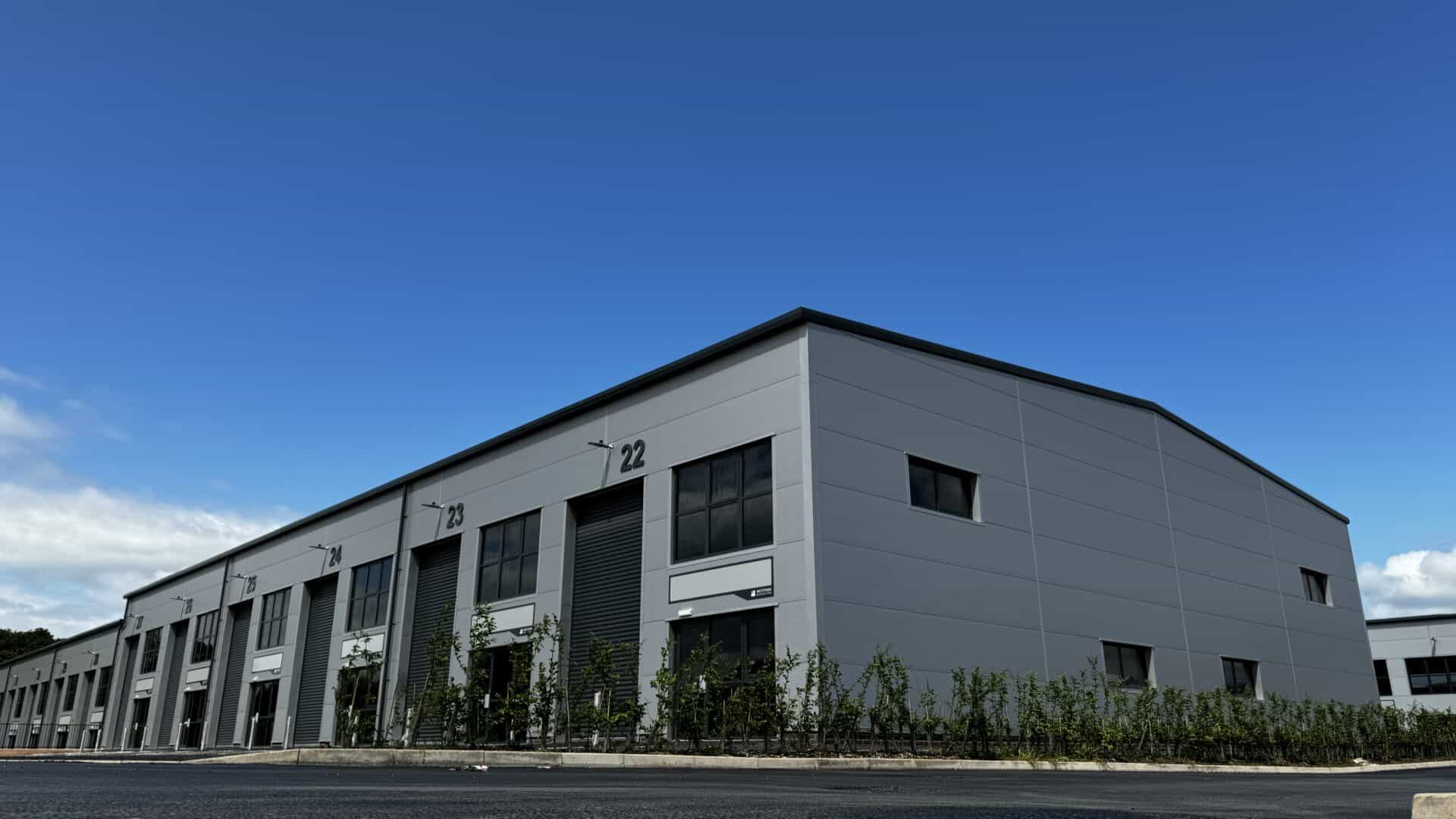 Exterior view of a modern industrial building under a clear blue sky. The building features large windows and multiple unit entrances, with a clean, grey facade. Young shrubs are planted along the perimeter.