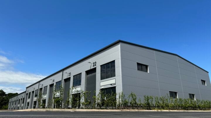 A modern industrial building with a sleek gray facade, large numbered units (22, 23, and 24), and dark glass windows. The building is surrounded by young green shrubs, set against a bright blue sky.