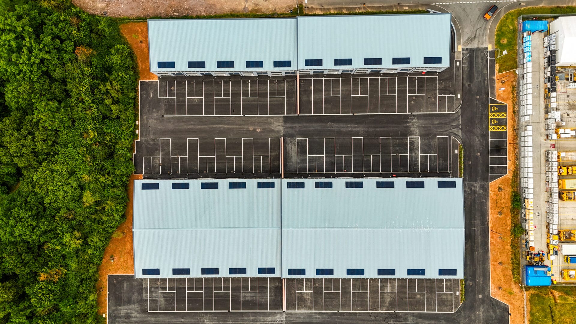 An overhead view of a commercial development featuring two large, light blue-roofed buildings surrounded by paved parking lots with marked spaces. To the left, there is a dense area of green trees, while the right side includes construction materials and vehicles. The layout is symmetrical and organized, with clear roadways connecting the site.