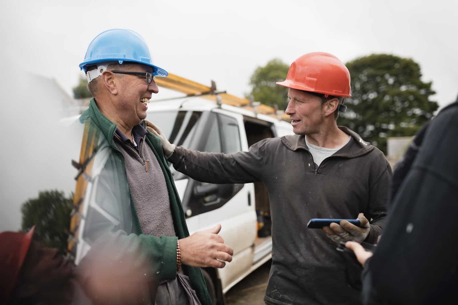 Group of blue collar workers standing outside while taking a break from work. They're all standing around, laughing and smiling, while looking at a smart phone.