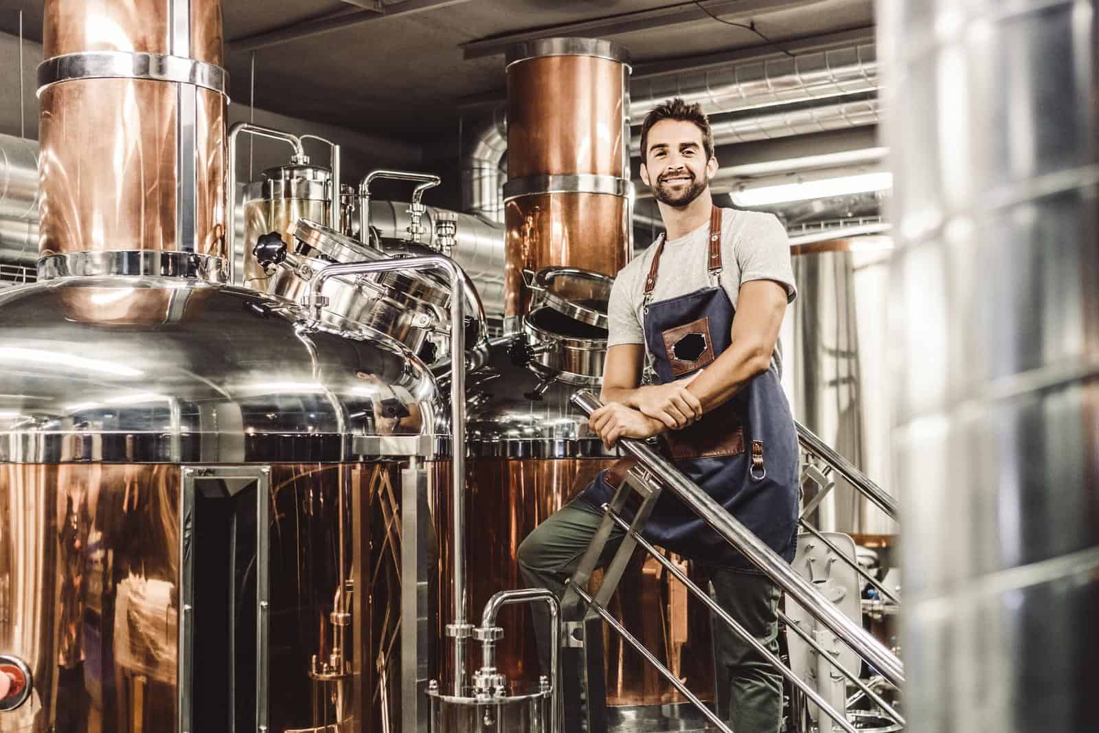 Low angle view of manager standing by distillery stills. Portrait of handsome male owner is on staircase at brewery. He is wearing apron.